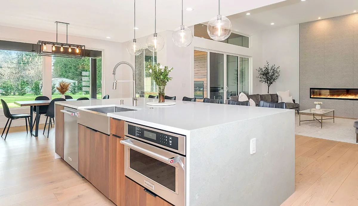 Open plan kitchen with a large island in light stone with a waterfall end, wood cabinet fronts and a living room with a linear fireplace behind
