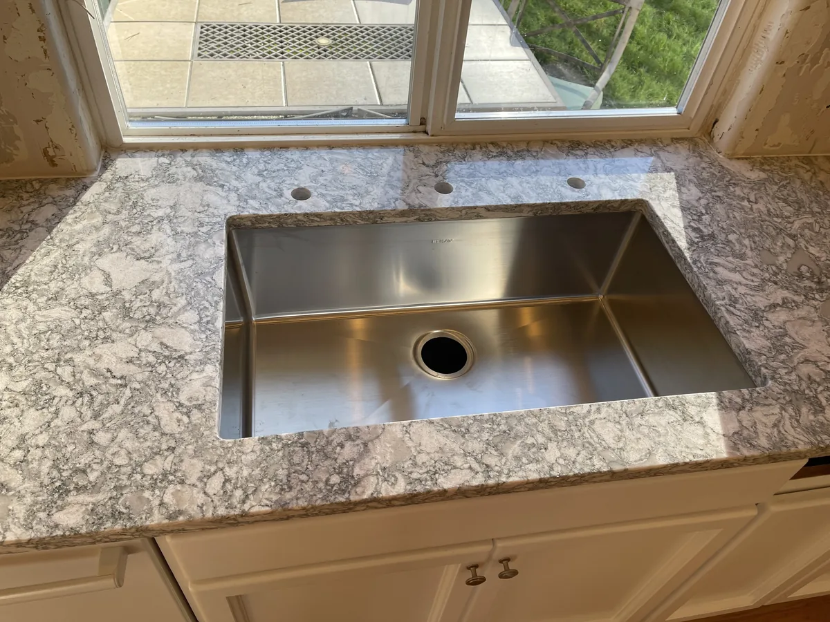 Undermount stainless sink set into a gray veined quartz countertop under a kitchen window, with drilled faucet holes