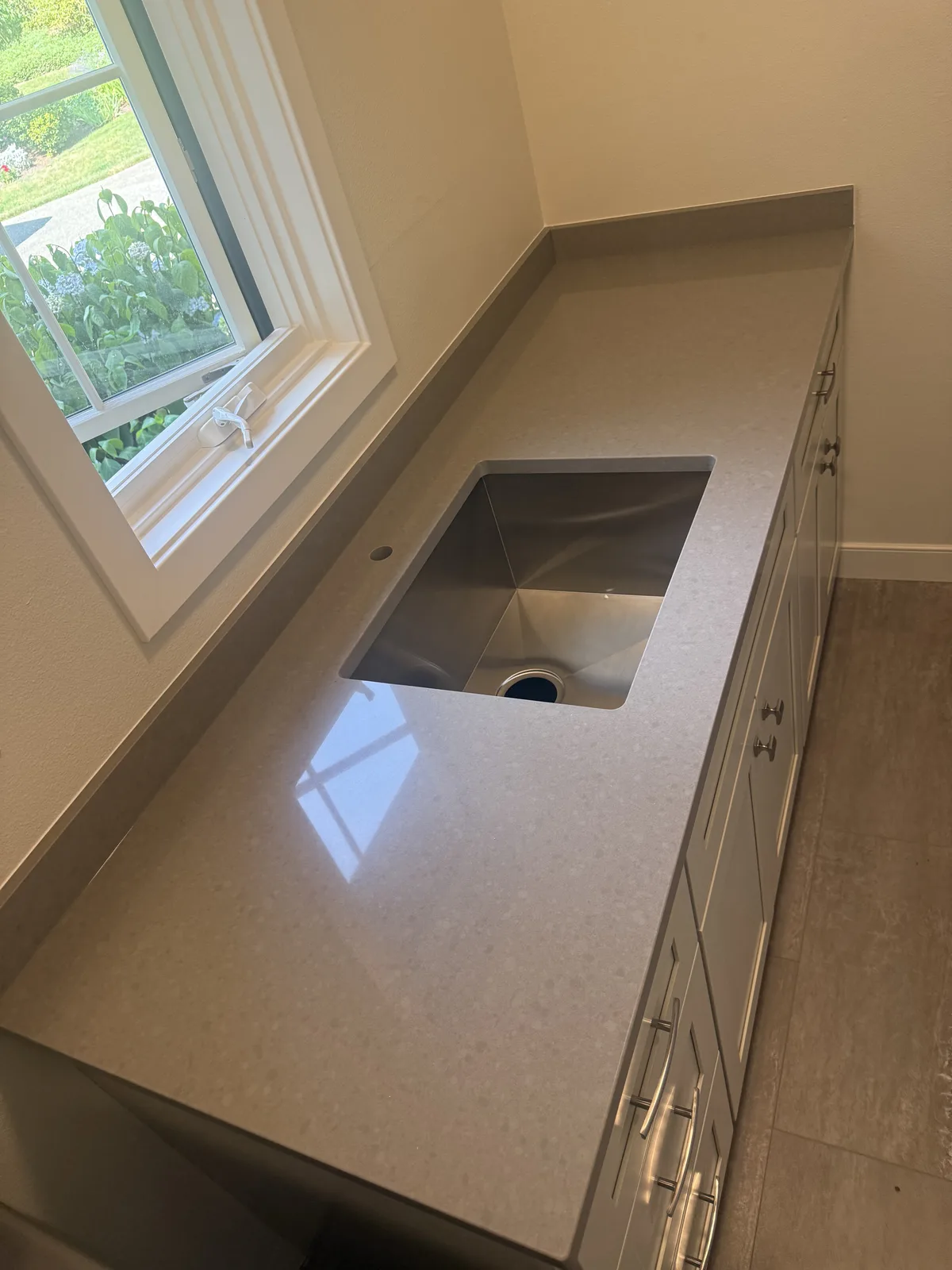 Gray engineered stone countertop with an undermount stainless sink, a drilled faucet hole, a short matching backsplash and a window above the run