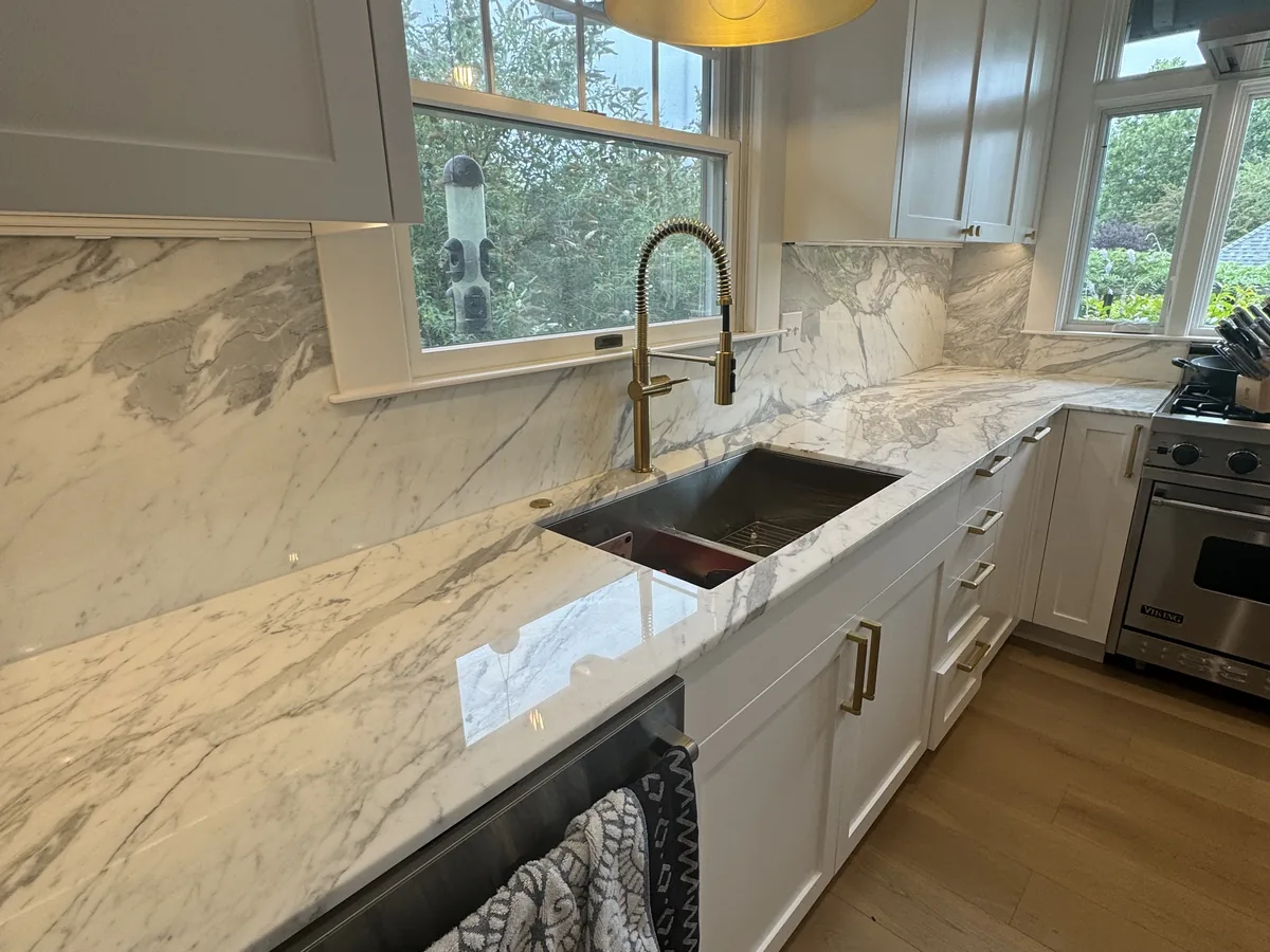 Veined stone countertop with a matching full height backsplash, an undermount workstation sink and a gold faucet under a kitchen window