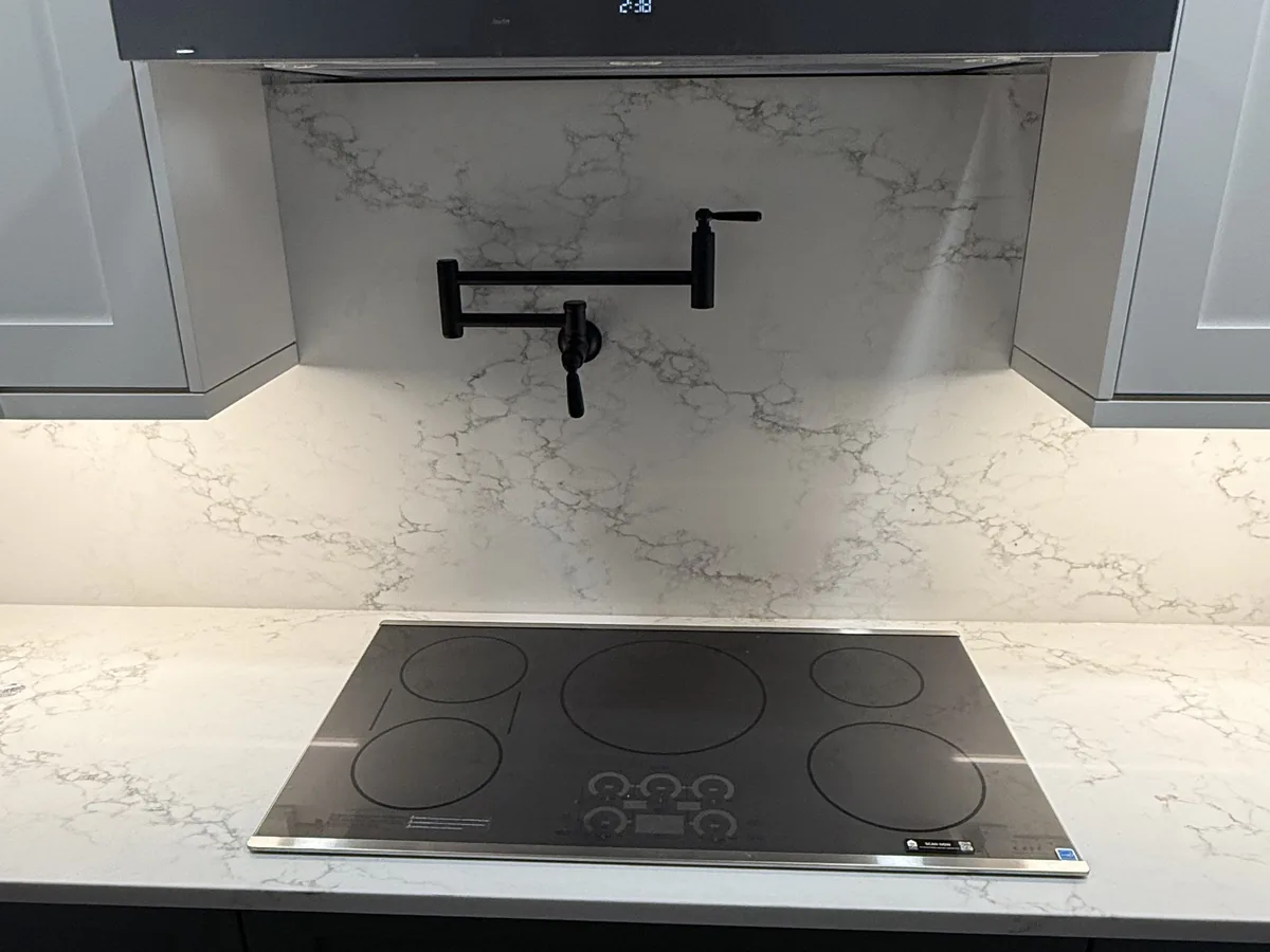 Induction cooktop set into a white veined quartz countertop with a matching full height quartz backsplash and a black pot filler