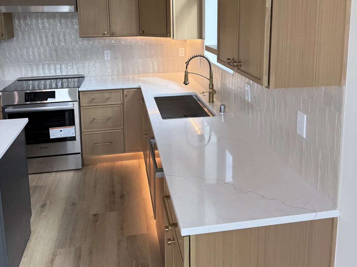 Quartz countertop with an undermount sink and a full tile backsplash, beside a stainless range in a light wood kitchen
