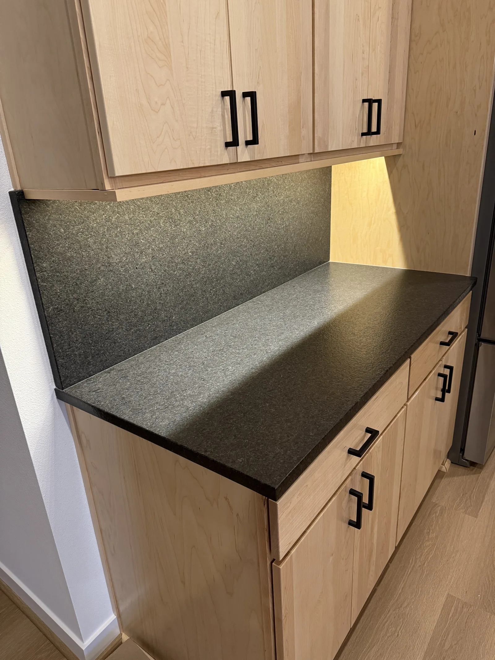 Black stone bar top with a full-height backsplash and light wood cabinets